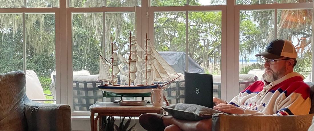 Shane Yerkes writing on a laptop in a sunroom, live oaks and Spanish moss through the windows behind him