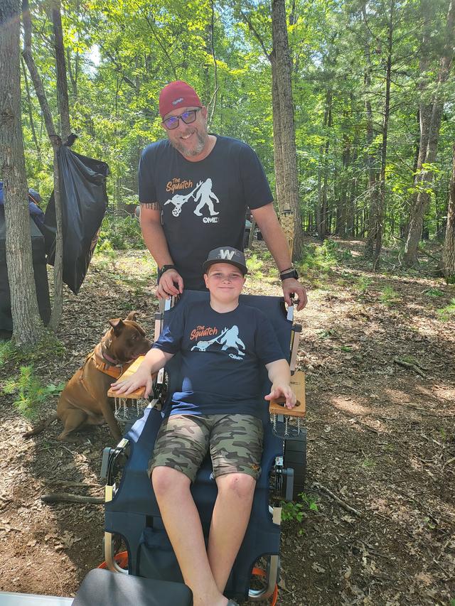 Shane and Conor riding a utility vehicle down a wooded trail with their dog alongside