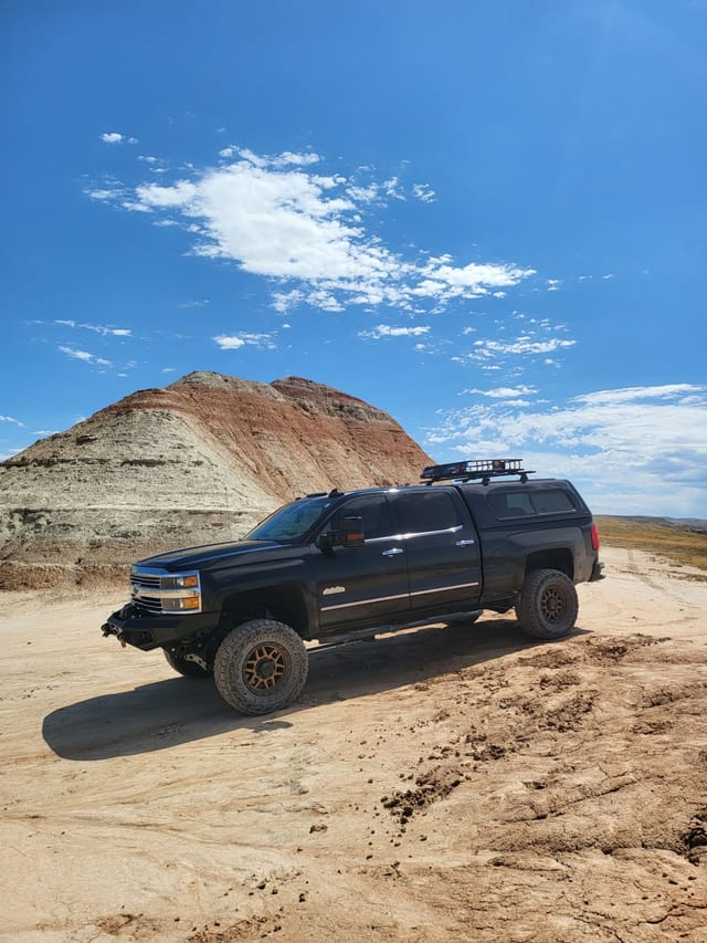 The black overland truck parked on cracked dirt beneath a striped Badlands hill under a big sky