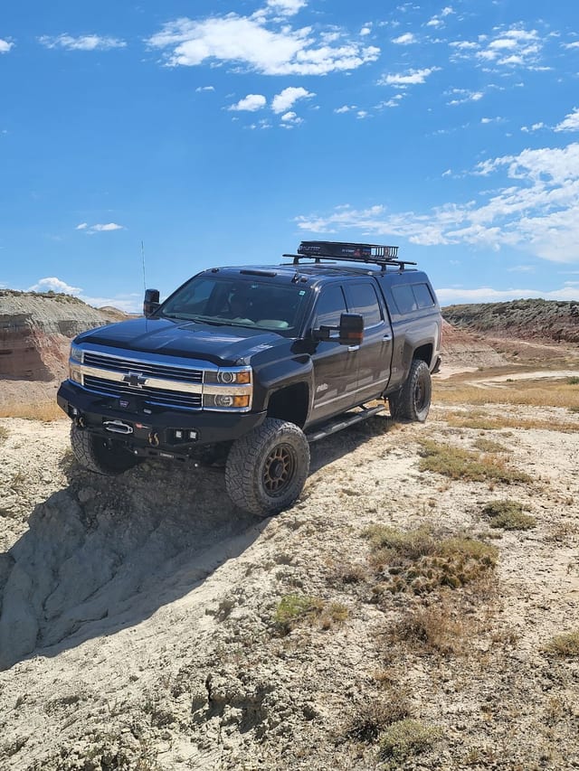 The black overland truck nosing over a pale ledge in the Badlands under a blue sky