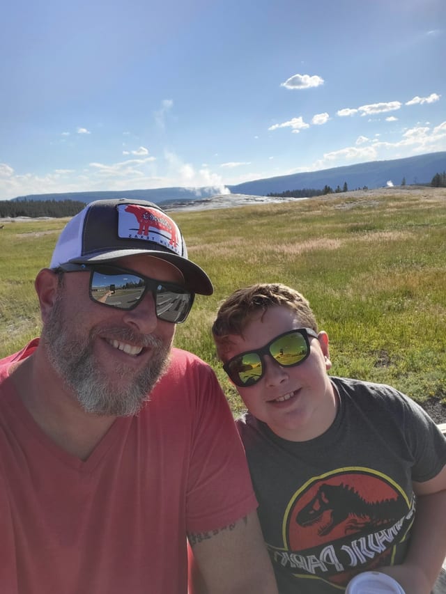 Shane and Conor in sunglasses, grinning, Old Faithful steaming on the horizon behind them