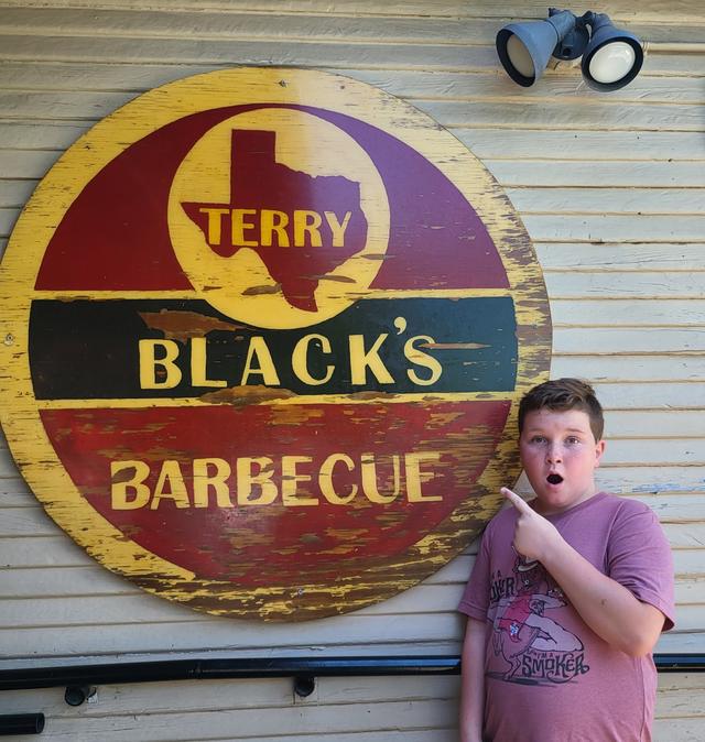 Conor pointing up at a large painted Terry Black's Barbecue sign, mouth open in delight