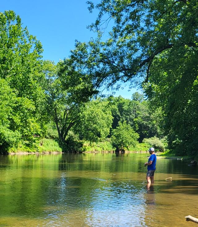 A boy standing alone in a wide, shallow green river lined with trees