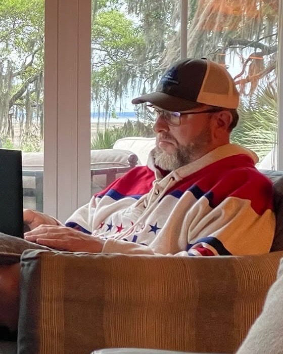 Shane Yerkes writing on a laptop in a sunroom, live oaks and Spanish moss through the windows behind him
