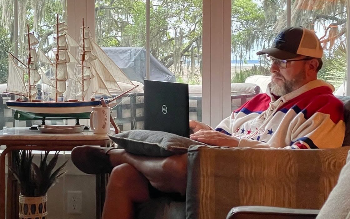 Shane Yerkes writing on a laptop in a sunroom, live oaks and Spanish moss through the windows behind him